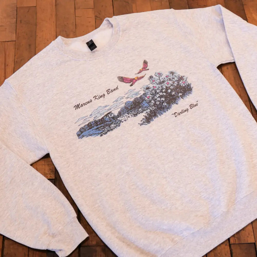 Heather gray long-sleeved sweatshirt featuring a colorful graphic of two birds flying over a landscape with flowers and text reading ’marcus king band’ and ’’darling blue’’.