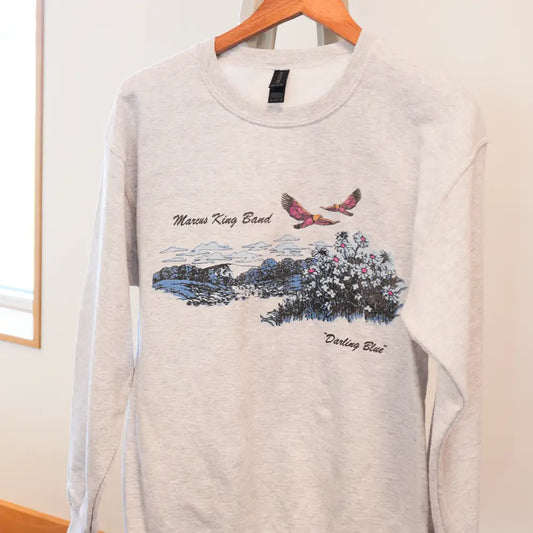 A heather gray long-sleeve sweatshirt featuring a printed design of two pink birds flying over a landscape with flowers and the text ’marcus king band’ above and ’’darling blue’’ below.