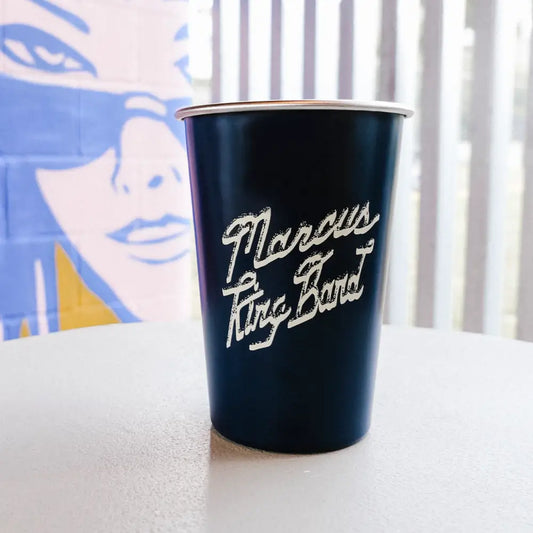 Navy blue paper cup with silver rim and white cursive ’marcus king band’ logo