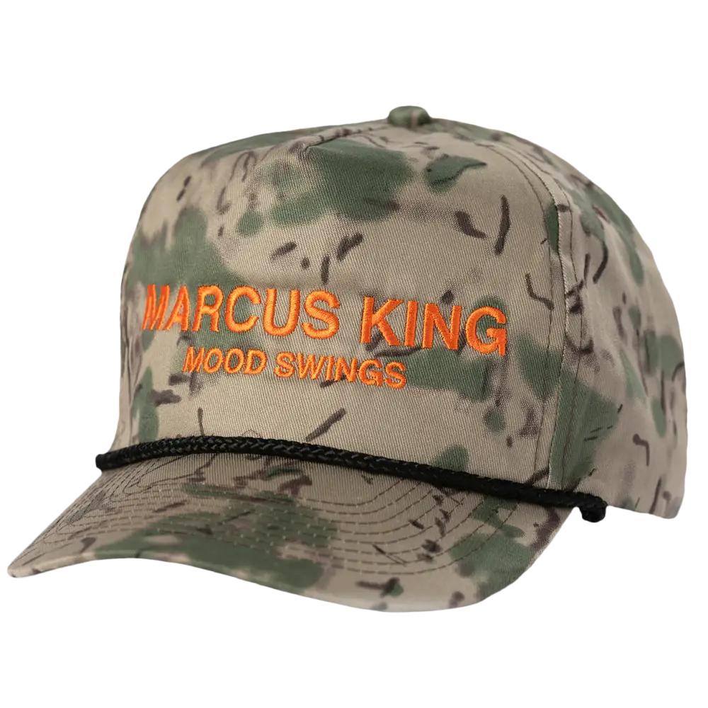 A camouflage-patterned baseball cap with orange embroidered text reading ’marcus king’ and ’wood swings,’ featuring a black braided cord around the brim.