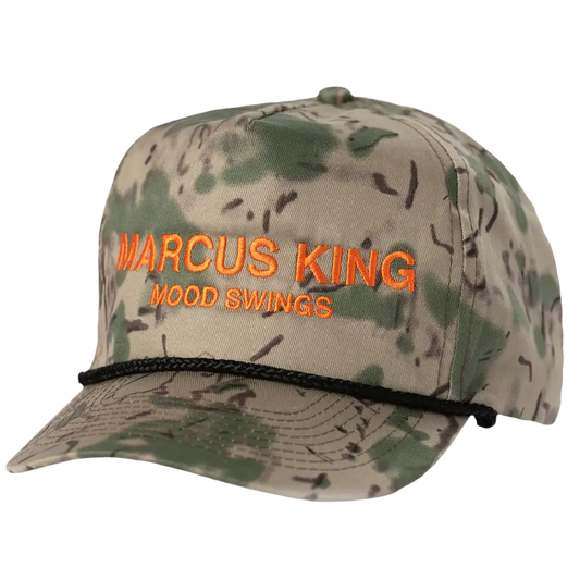 A camouflage-patterned baseball cap with orange embroidered text reading ’marcus king’ and ’wood swings,’ featuring a black braided cord around the brim.